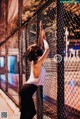 A woman in a white tank top and black leggings leaning against a fence.