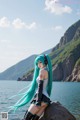 A woman with long blue hair sitting on a rock by the water.